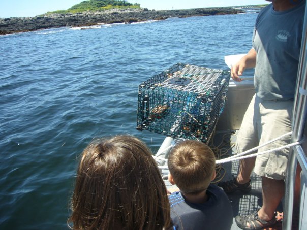 two children looking at a lobster trap in Maine, a person standing to the right holding the lobster trap